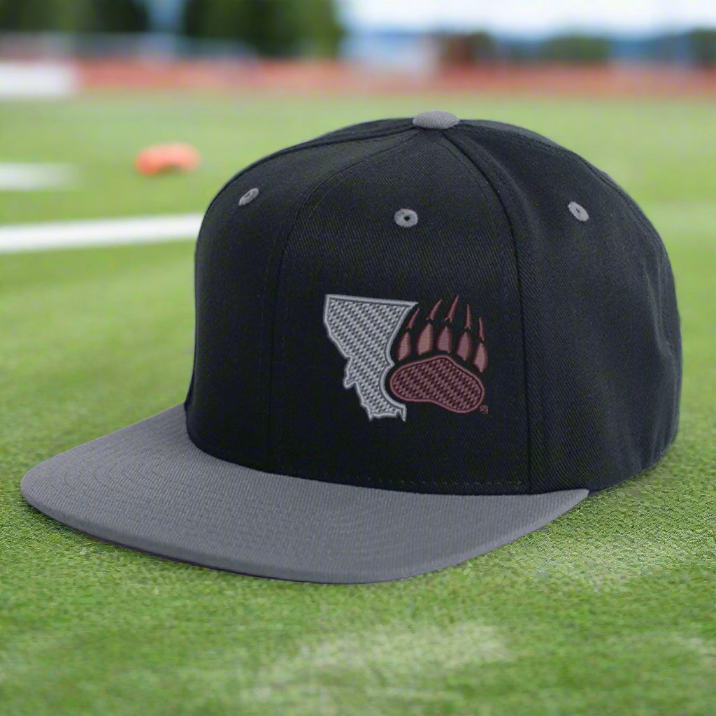 Black Montana Paw - Wool Flat Bill Hat made in Missoula, MT (home of the UM Grizzlies) by Blue Peak Creative—a black flat bill hat embroidered with a bear paw and the western edge of the state of Montana, sitting on a football field.