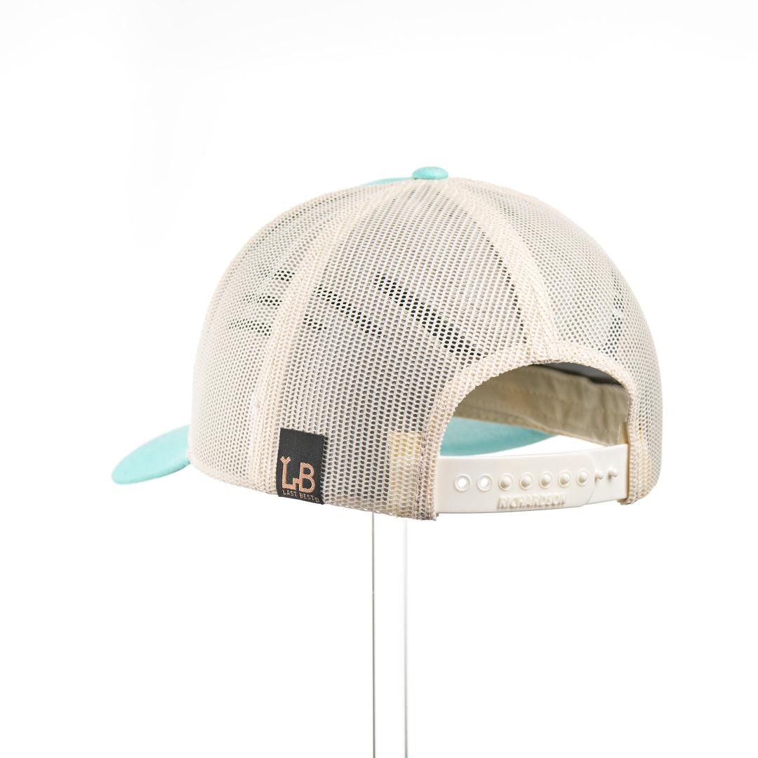 The back of a Cherry Wood Steer Skull w/ Paisley Teal & Cream Trucker Hat, featuring a hexagonal real cherry wood veneer patch with cut-out steer skull silhouette surrounded by etched paisley design, laminated to a hexagonal teal anodized aluminum plate, and riveted to a light teal and cream Richardson 115 low-profile trucker hat—handmade in Montana, USA by Last Best Supply Co.