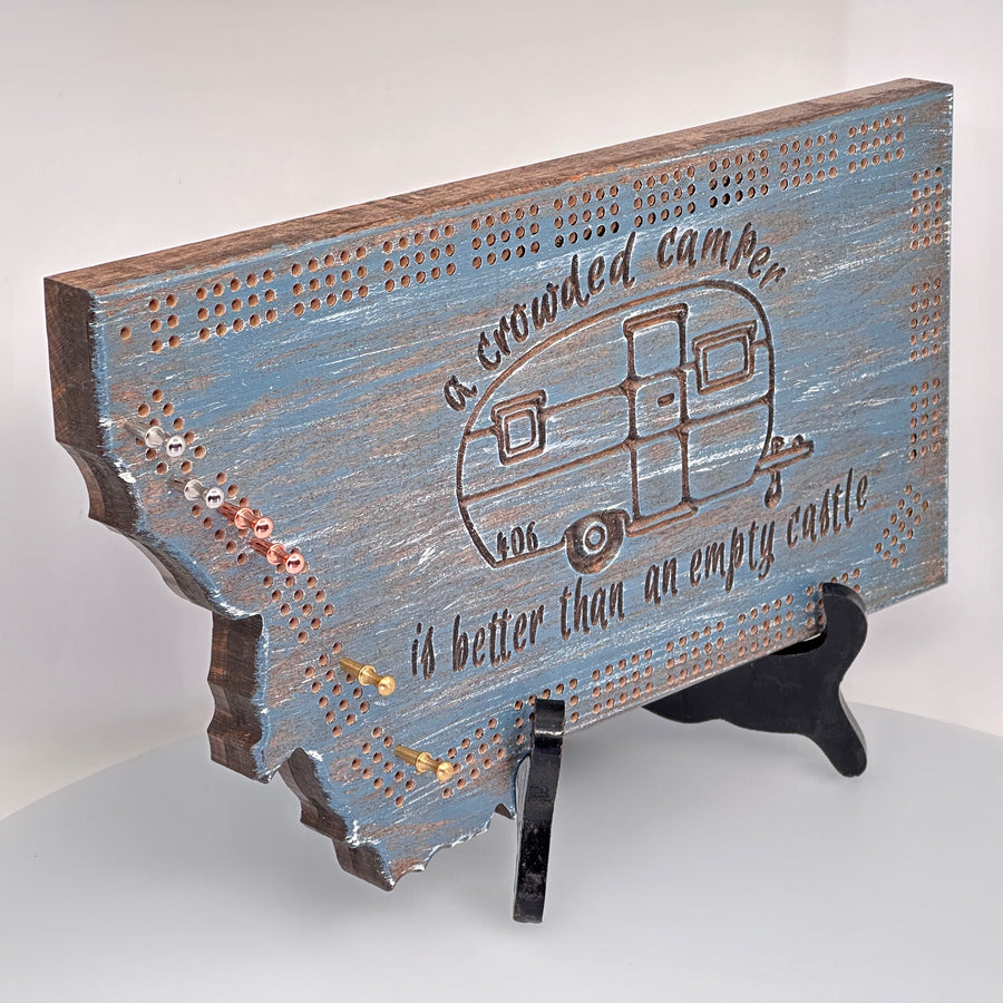 Knotty Pine Woodworks Handmade Camper Cribbage Board Lakeside (front)--this made in Montana wooden cribbage board is shaped like the outline of the state of Montana, painted in the 'lakeside' color scheme,  and features a camping-themed engraved design with a vintage camper and the expression, 'a crowded camper is better than an empty castle.'