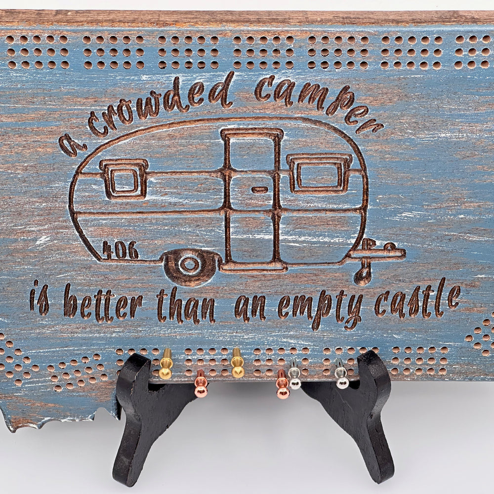 Knotty Pine Woodworks Handmade Camper Cribbage Board Lakeside (front)--this made in Montana wooden cribbage board is shaped like the outline of the state of Montana, painted in the 'lakeside' color scheme,  and features a camping-themed engraved design with a vintage camper and the expression, 'a crowded camper is better than an empty castle.'