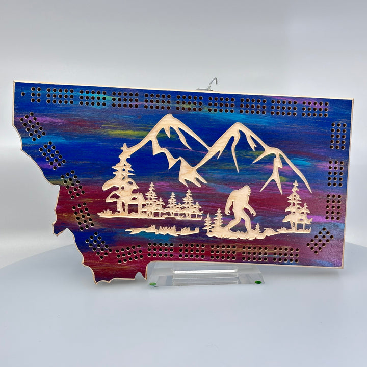 Knotty Pine Woodworks Handmade Montana Sasquatch Cribbage Board Sunset (front)--this made in Montana wooden cribbage board is shaped like the outline of the state of Montana, in sunset tones, and features an engraved Sasquatch striding in front of mountain peaks on the front.