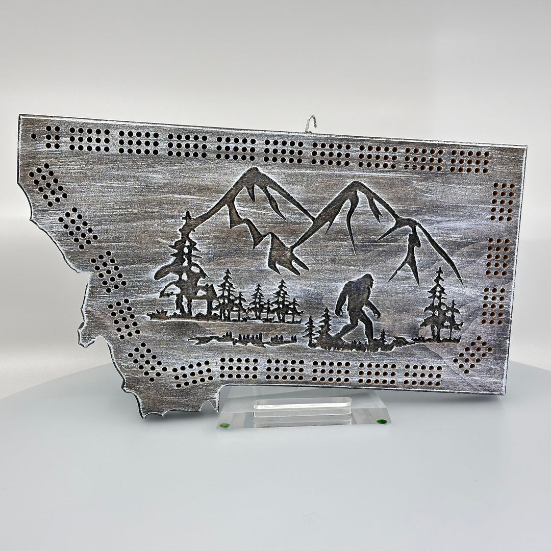 Knotty Pine Woodworks Handmade Montana Sasquatch Cribbage Board White/ Dark (front)--this made in Montana wooden cribbage board is shaped like the outline of the state of Montana, in weathered dark and light tones, and features an engraved Sasquatch striding in front of mountain peaks on the front.