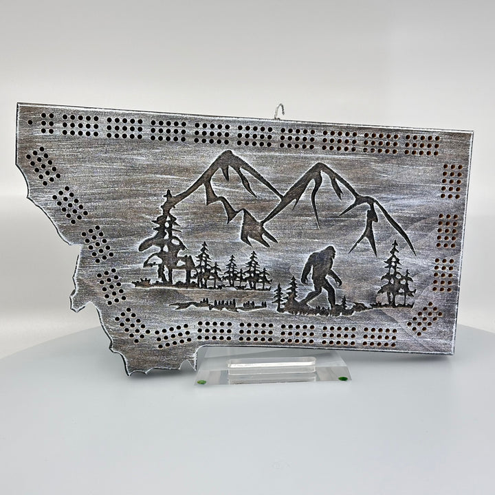 Knotty Pine Woodworks Handmade Montana Sasquatch Cribbage Board White/ Dark (front)--this made in Montana wooden cribbage board is shaped like the outline of the state of Montana, in weathered dark and light tones, and features an engraved Sasquatch striding in front of mountain peaks on the front.