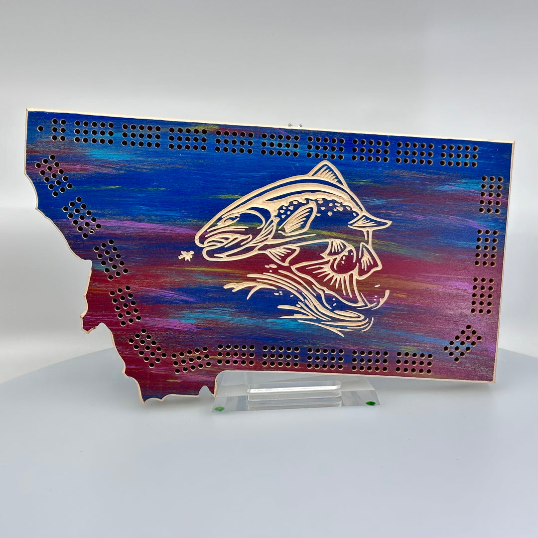 Knotty Pine Woodworks Handmade Montana Trout Cribbage Board Sunset (front)--this made in Montana wooden cribbage board is shaped like the outline of the state of Montana, in sunset tones, and features an engraved trout jumping out of water on the front.