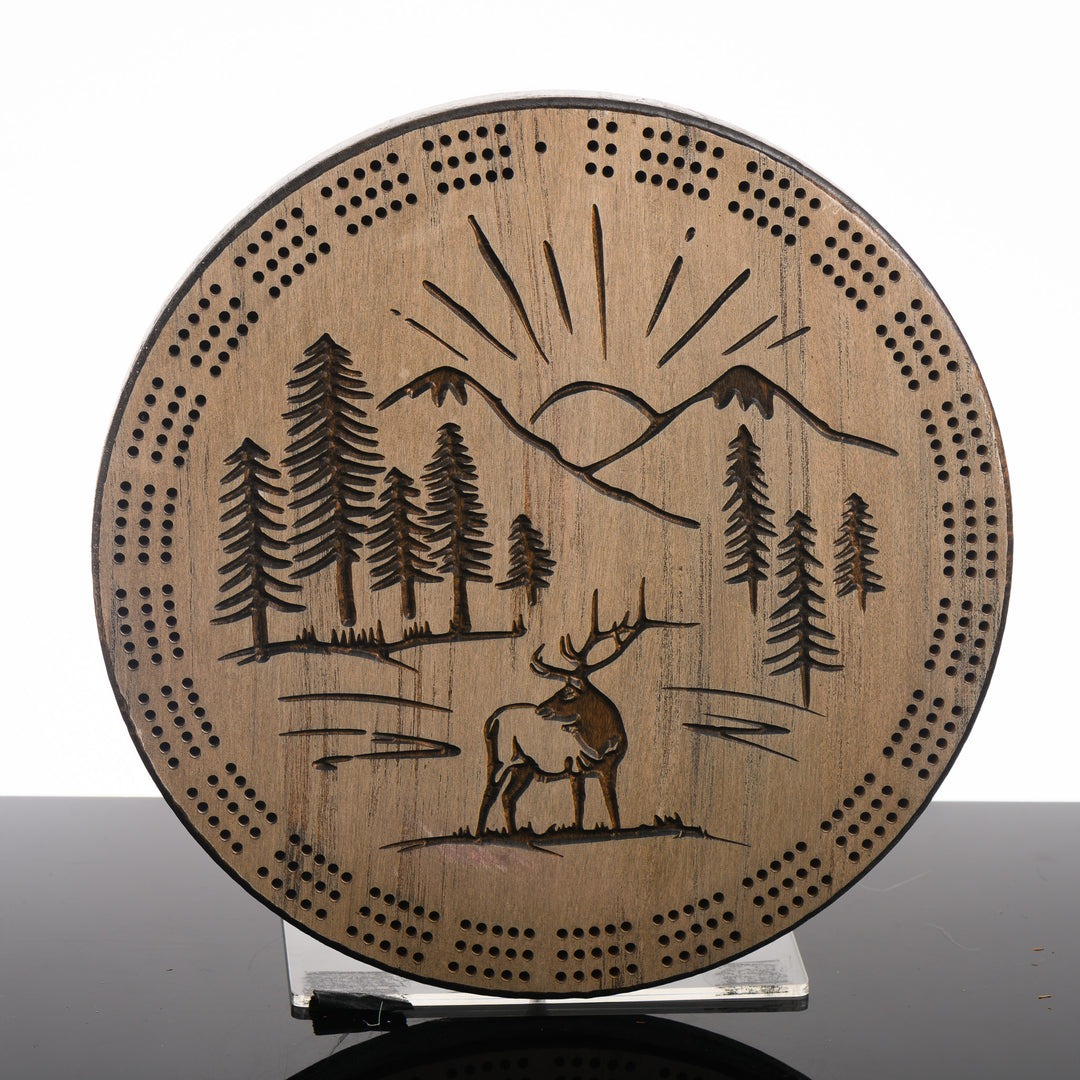 Knotty Pine Woodworks Handmade Mountain Elk Circle Cribbage Board Barn Wood Dark (front)--this made in Montana wooden cribbage board is circular in shape with a barn wood finish and dark details, and features a beautifully engraved elk standing proudly against a backdrop of sunbeams framed by mountain peaks on the front.