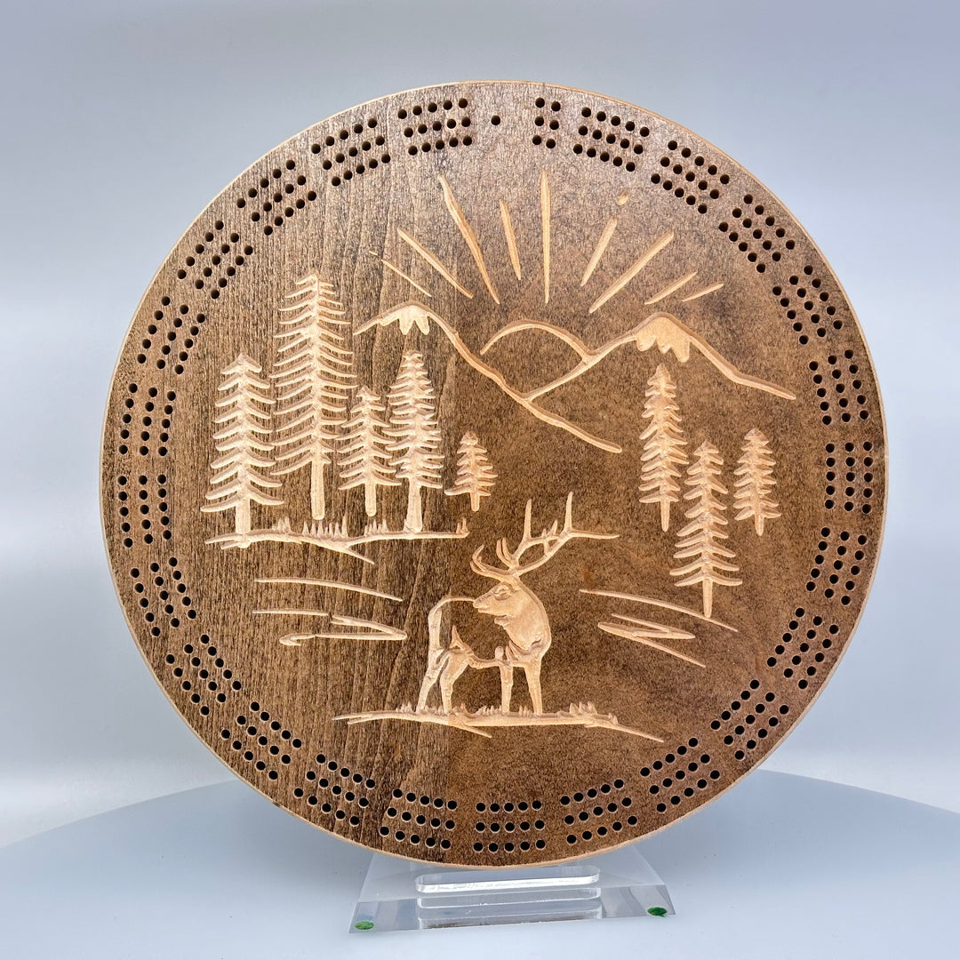 Knotty Pine Woodworks Handmade Mountain Elk Circle Cribbage Board Light Espresso (front)--this made in Montana wooden cribbage board is circular in shape with a light espresso finish, and features a beautifully engraved elk standing proudly against a backdrop of sunbeams framed by mountain peaks on the front.