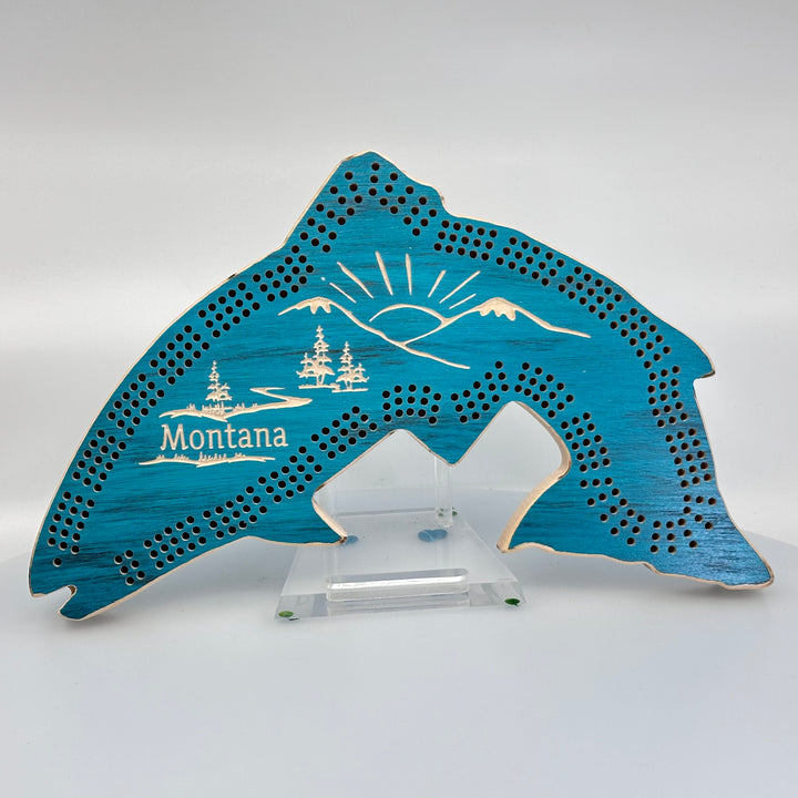 Knotty Pine Woodworks Handmade Trout Cribbage Board with Montana Mountain Scene Turquoise (front)--this made in Montana wooden cribbage board is shaped like a leaping trout, finished in turquoise, and features a beautifully engraved forest and mountain scene with the word ‘Montana’ engraved on the front.