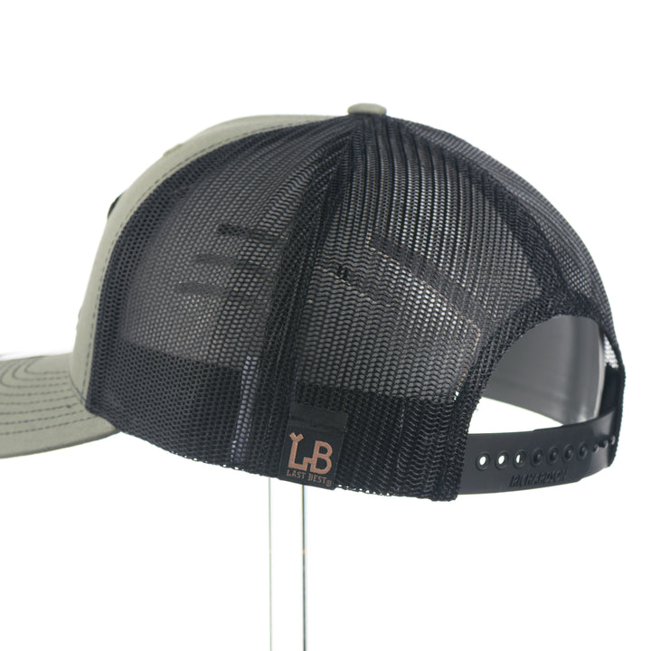 Back of a Montana Walnut Wood with Sundae Copper Patch on Richardson Trucker Hat, featuring a rectangular, real walnut wood veneer patch with a precision-cut state of Montana silhouette cutout, laminated to a black anodized metal plate, and then riveted to an olive green Richardson 112 trucker hat with black mesh—handmade in Montana, USA by Last Best Supply Co.