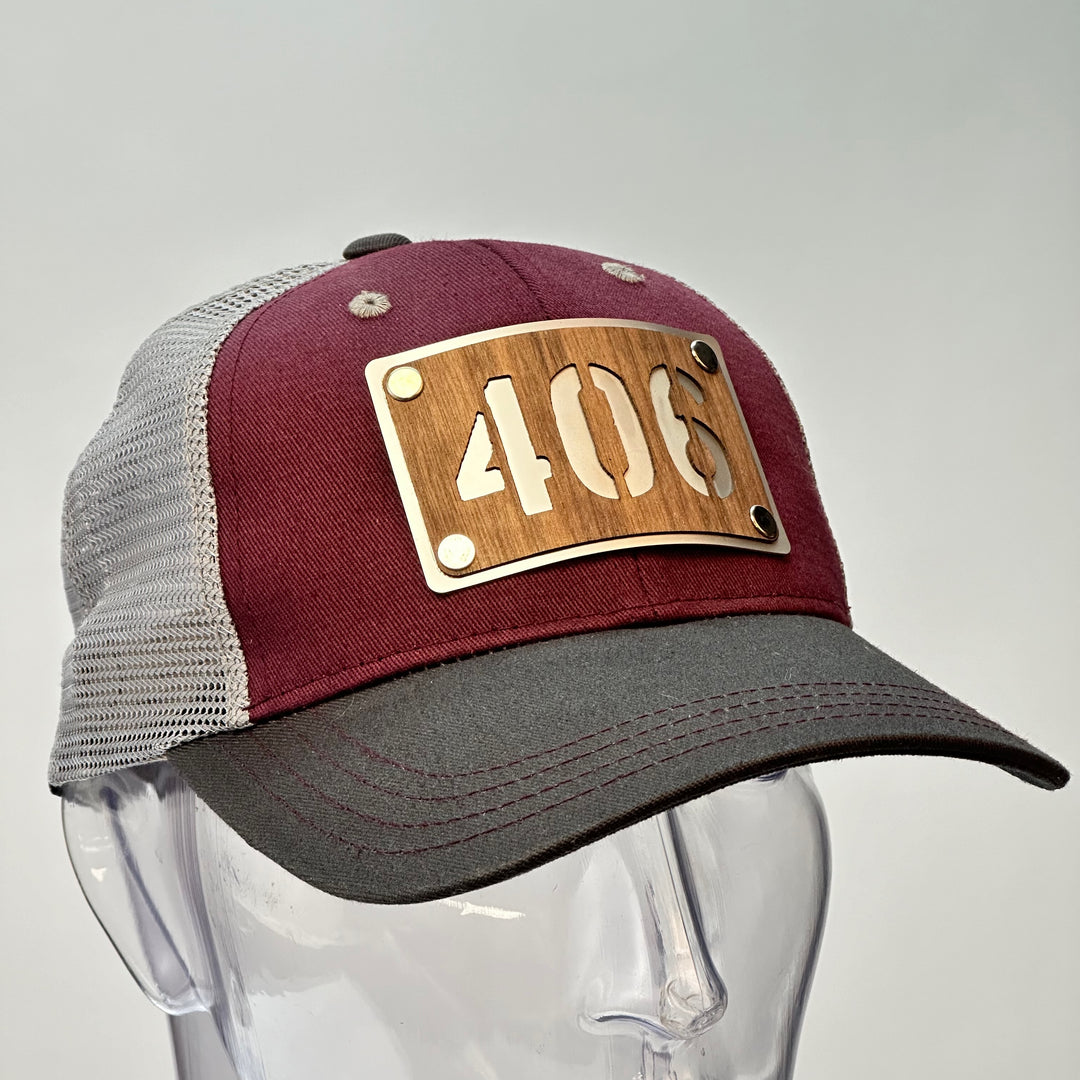 3/4 of a 406 Walnut Wood & Metal Patch Maroon, Charcoal and Grey Trucker Hat, featuring a rectangular, real cherry wood veneer patch with a precision-cut “406” cutout, laminated to a silver anodized metal plate, and then riveted to a maroon and charcoal grey mesh trucker hat—handmade in Montana, USA by Last Best Supply Co.