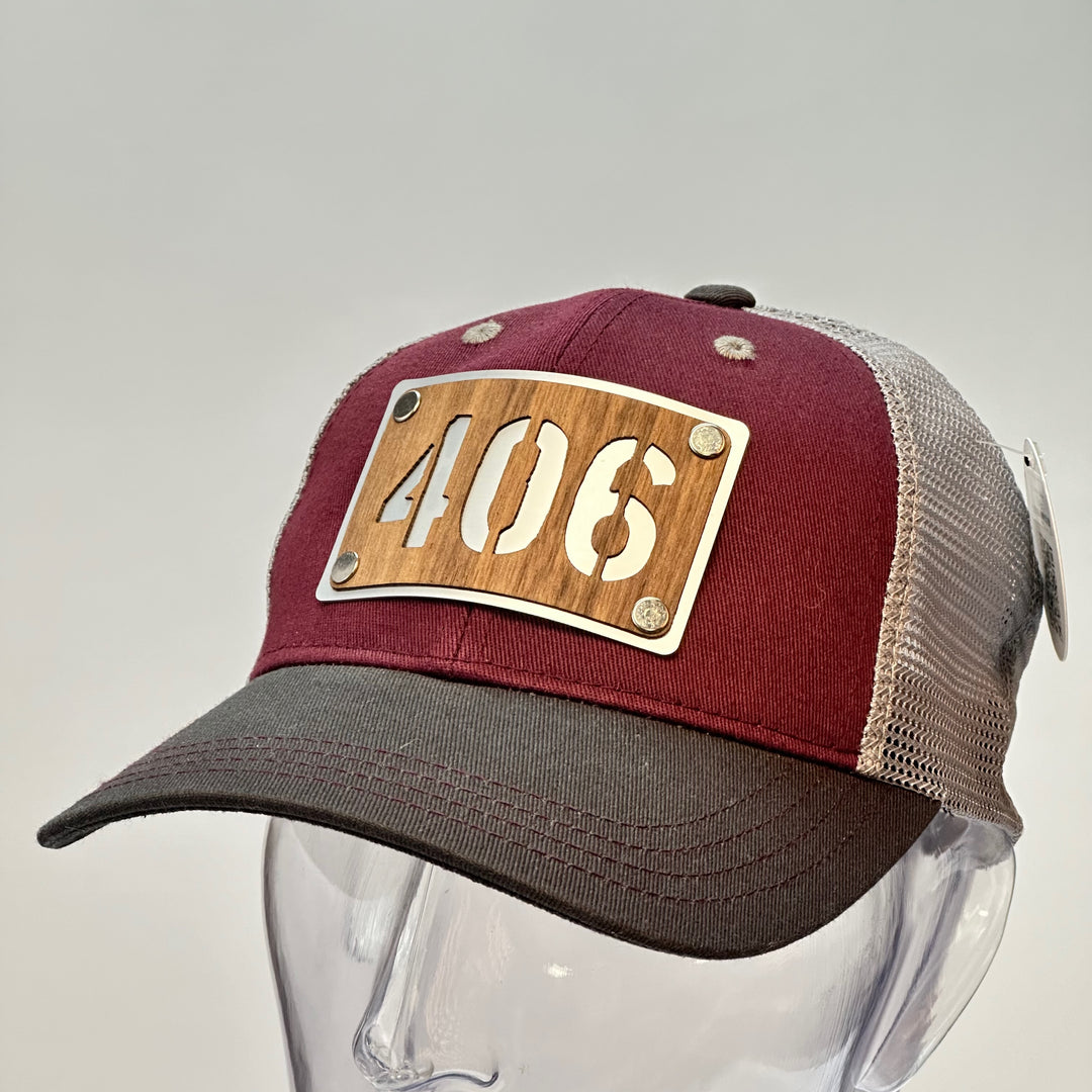 3/4 view of a 406 Walnut Wood & Metal Patch Maroon, Charcoal and Grey Trucker Hat, featuring a rectangular, real cherry wood veneer patch with a precision-cut “406” cutout, laminated to a silver anodized metal plate, and then riveted to a maroon and charcoal grey mesh trucker hat—handmade in Montana, USA by Last Best Supply Co.