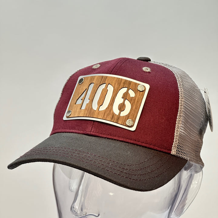 3/4 view of a 406 Walnut Wood & Metal Patch Maroon, Charcoal and Grey Trucker Hat, featuring a rectangular, real cherry wood veneer patch with a precision-cut “406” cutout, laminated to a silver anodized metal plate, and then riveted to a maroon and charcoal grey mesh trucker hat—handmade in Montana, USA by Last Best Supply Co.