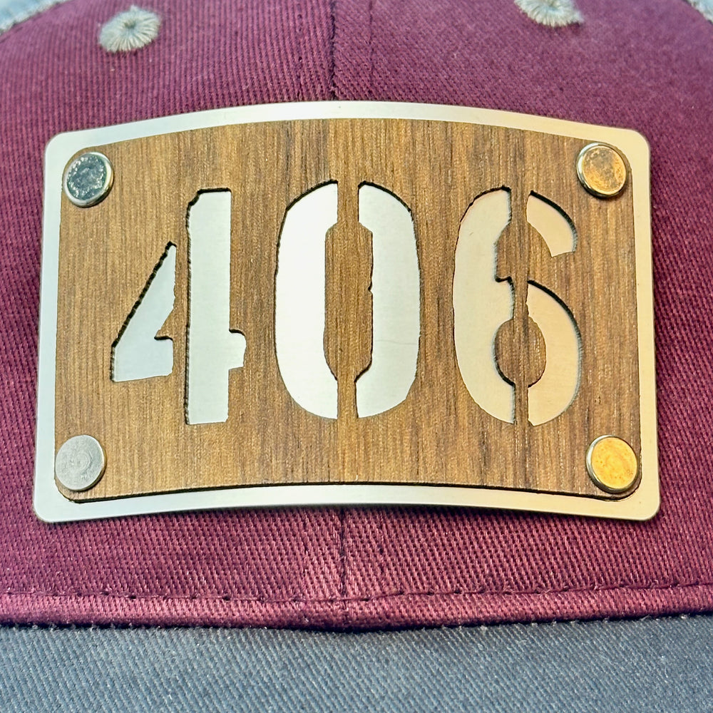 Patch detail of a 406 Walnut Wood & Metal Patch Maroon, Charcoal and Grey Trucker Hat, featuring a rectangular, real cherry wood veneer patch with a precision-cut “406” cutout, laminated to a silver anodized metal plate, and then riveted to a maroon and charcoal grey mesh trucker hat—handmade in Montana, USA by Last Best Supply Co.