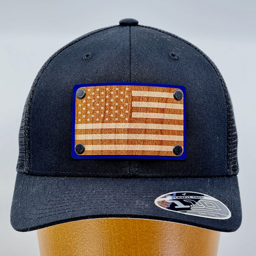 American Flag Wood & Blue Metal Patch Flexfit Trucker Hat (Black) featuring an American Flag etched on cherry wood veneer and laminated to a blue metal plate—handmade in Montana, USA by Last Best Supply Co. (front view)