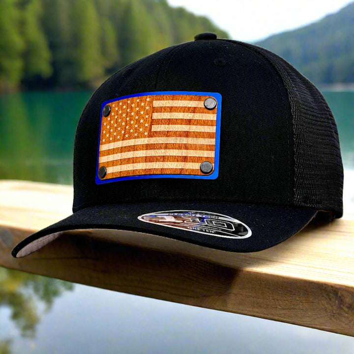 American Flag Wood & Blue Metal Patch Flexfit Trucker Hat (Black) featuring an American Flag etched on cherry wood veneer and laminated to a blue metal plate—handmade in Montana, USA by Last Best Supply Co. (3/4 view)