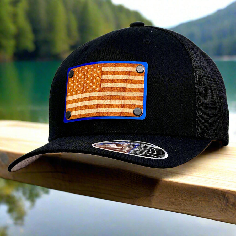 American Flag Wood & Blue Metal Patch Flexfit Trucker Hat (Black) featuring an American Flag etched on cherry wood veneer and laminated to a blue metal plate—handmade in Montana, USA by Last Best Supply Co. (3/4 view)