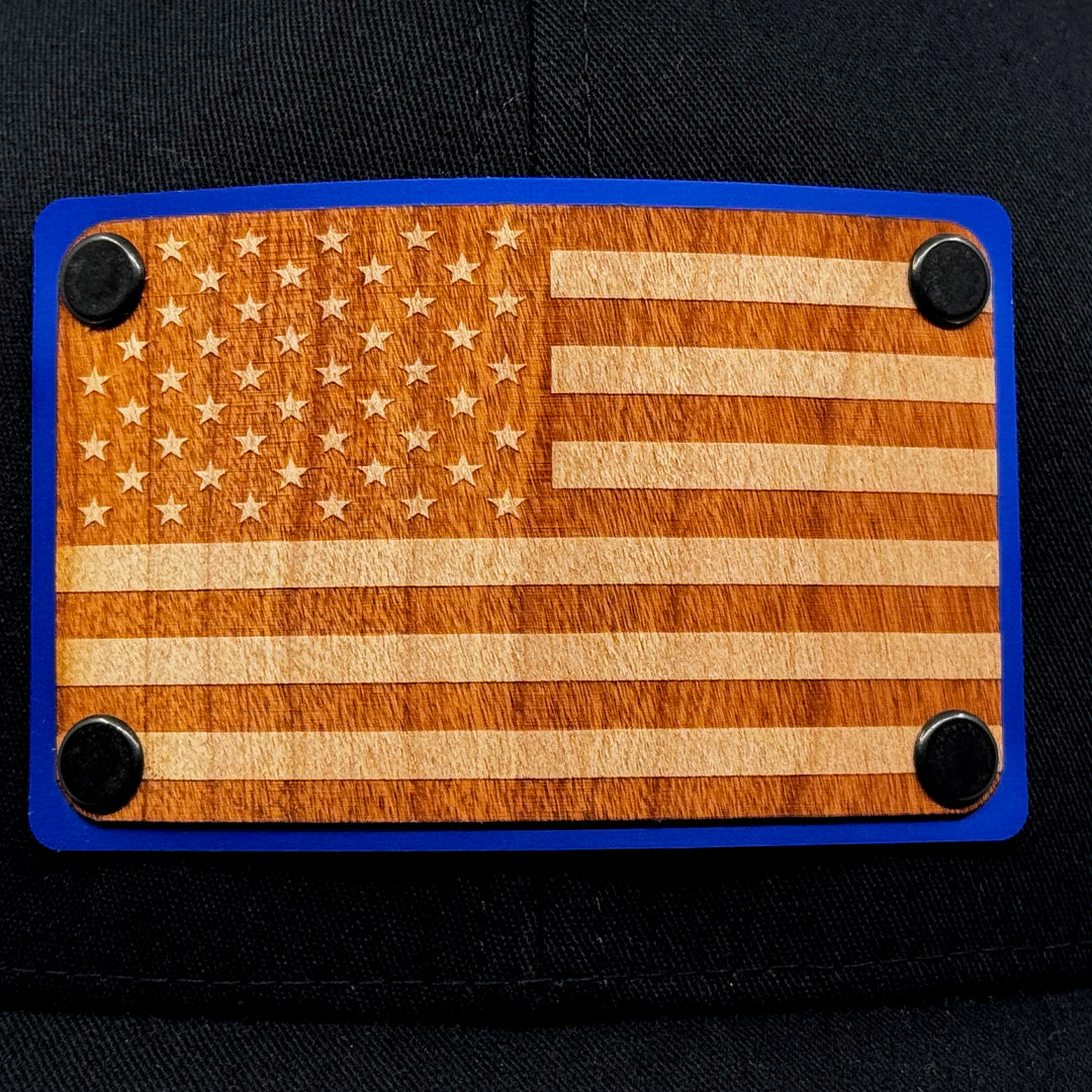 American Flag Wood & Blue Metal Patch Flexfit Trucker Hat (Black) featuring an American Flag etched on cherry wood veneer and laminated to a blue metal plate—handmade in Montana, USA by Last Best Supply Co. (patch detail)