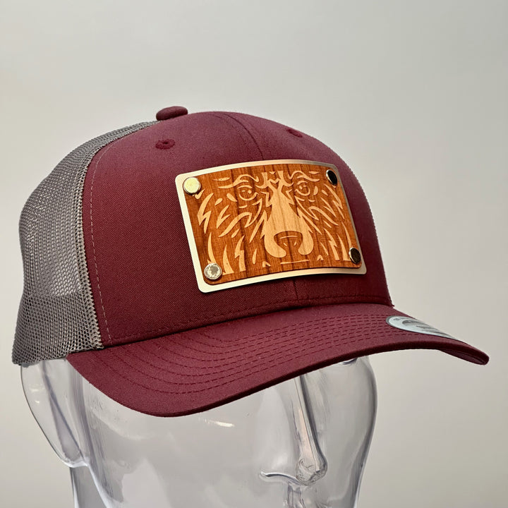 3/4 view of a Bear Face Cherry Wood & Silver Metal Patch Maroon and Grey Trucker Hat, featuring a rectangular, real cherry wood veneer patch etched with a bold bear face design, laminated to a silver anodized metal plate, and then riveted to a maroon and charcoal grey mesh trucker hat—handmade in Montana, USA by Last Best Supply Co.