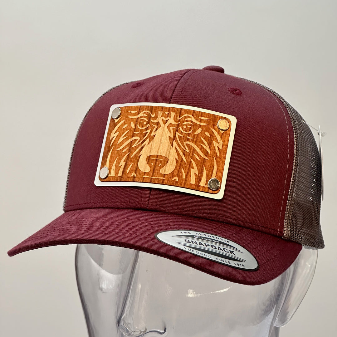 3/4 view of a Bear Face Cherry Wood & Silver Metal Patch Maroon and Grey Trucker Hat, featuring a rectangular, real cherry wood veneer patch etched with a bold bear face design, laminated to a silver anodized metal plate, and then riveted to a maroon and charcoal grey mesh trucker hat—handmade in Montana, USA by Last Best Supply Co.