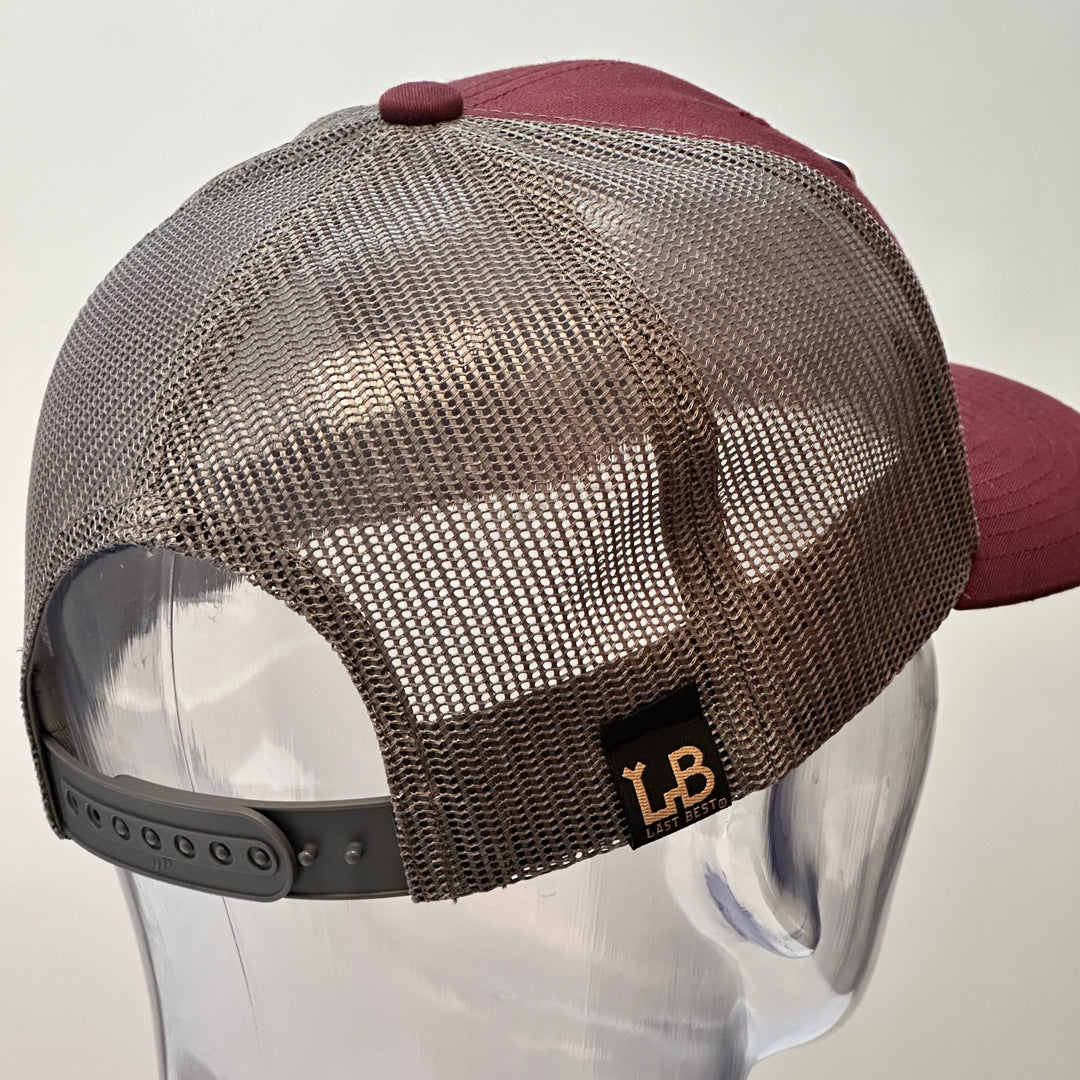 The back of a Bear Face Cherry Wood & Silver Metal Patch Maroon and Grey Trucker Hat, featuring a rectangular, real cherry wood veneer patch etched with a bold bear face design, laminated to a silver anodized metal plate, and then riveted to a maroon and charcoal grey mesh trucker hat—handmade in Montana, USA by Last Best Supply Co.