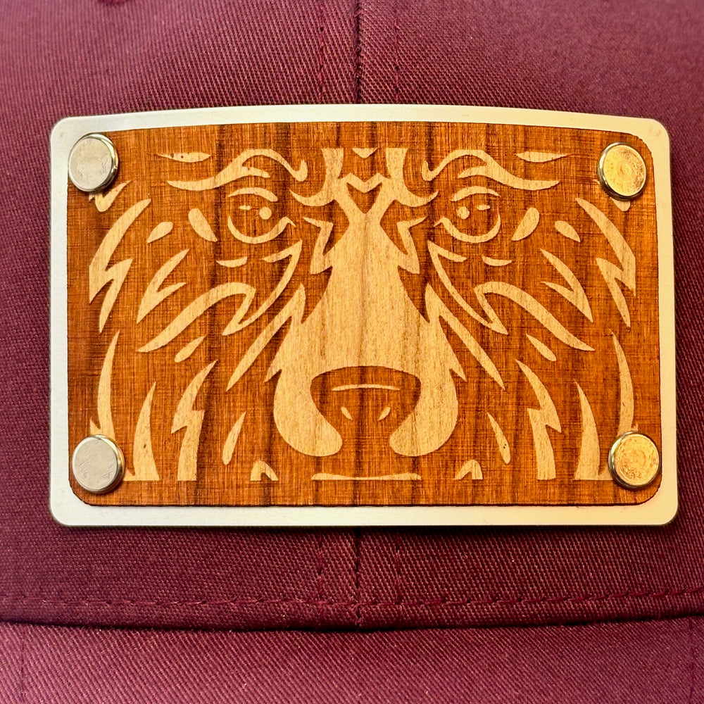 Patch detail of a Bear Face Cherry Wood & Silver Metal Patch Maroon and Grey Trucker Hat, featuring a rectangular, real cherry wood veneer patch etched with a bold bear face design, laminated to a silver anodized metal plate, and then riveted to a maroon and charcoal grey mesh trucker hat—handmade in Montana, USA by Last Best Supply Co.