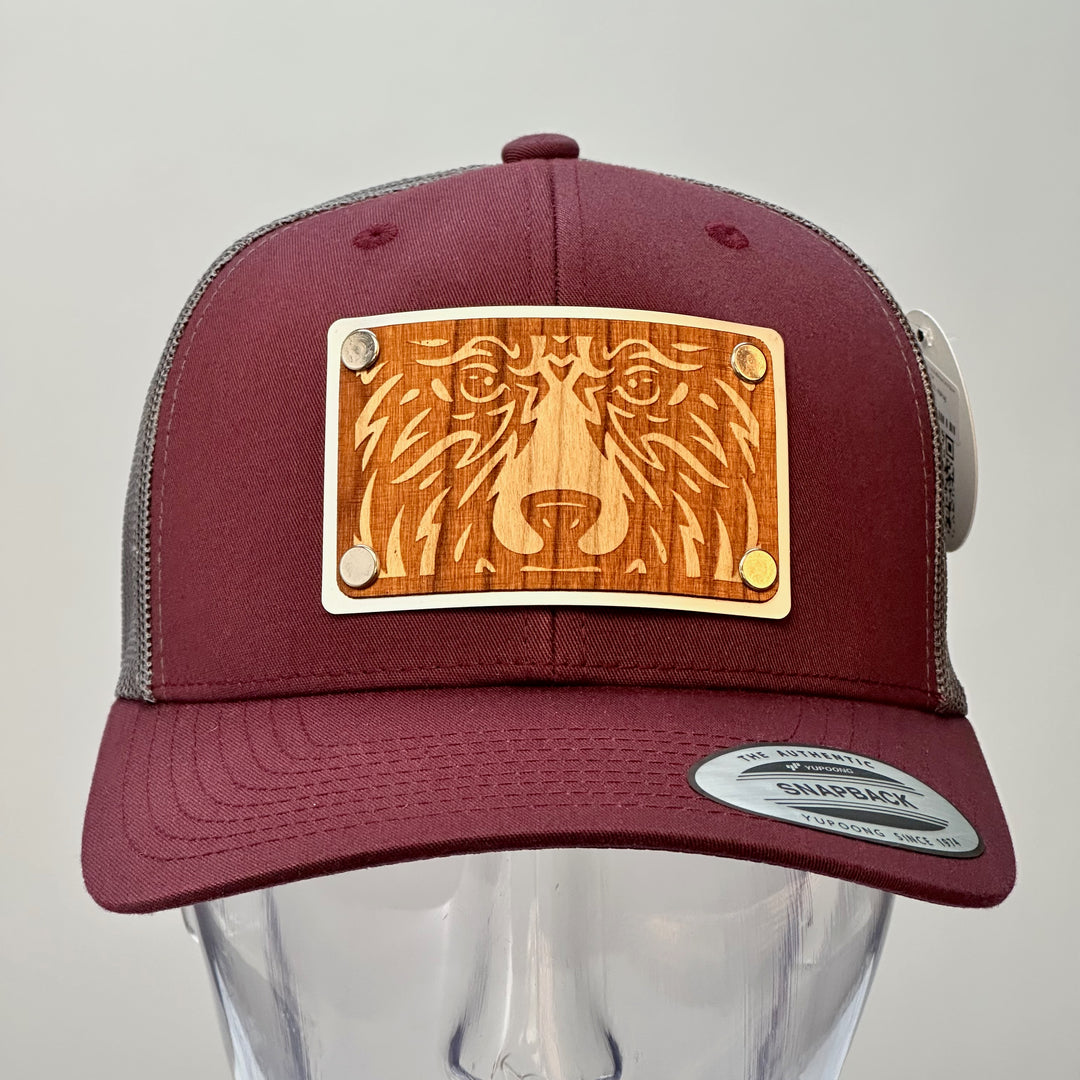 A front view of a Bear Face Cherry Wood & Silver Metal Patch Maroon and Grey Trucker Hat, featuring a rectangular, real cherry wood veneer patch etched with a bold bear face design, laminated to a silver anodized metal plate, and then riveted to a maroon and charcoal grey mesh trucker hat—handmade in Montana, USA by Last Best Supply Co.