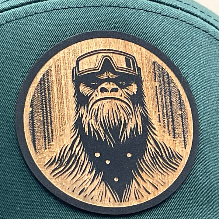 Patch detail of a Bigfoot Face Flexwood Patch Forest Green Flat Bill Trucker Hat, featuring a circular Flexwood patch etched with a Bigfoot face design and securely attached to a forest green and black Richardson flat bill trucker hat—handmade in Montana, USA by Last Best Supply Co.