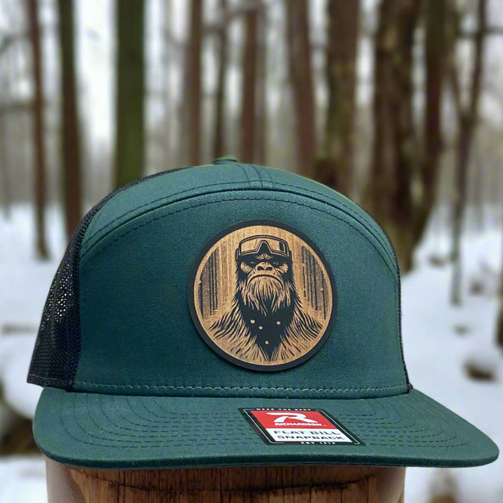Front of a Bigfoot Face Flexwood Patch Forest Green Flat Bill Trucker Hat, featuring a circular Flexwood patch etched with a Bigfoot face design and securely attached to a forest green and black Richardson flat bill trucker hat—handmade in Montana, USA by Last Best Supply Co.