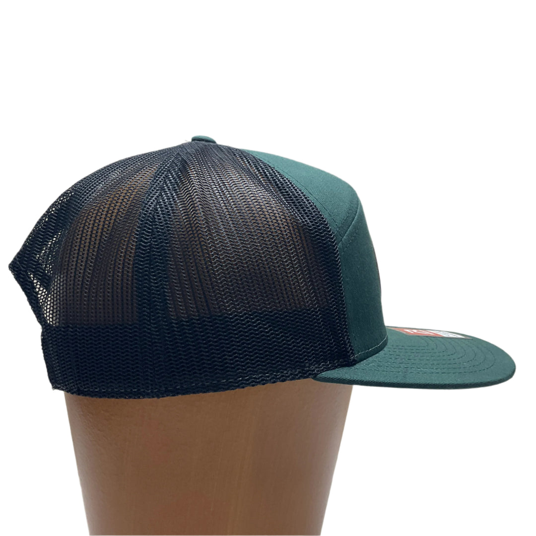 Side view of a Bigfoot Face Flexwood Patch Forest Green Flat Bill Trucker Hat, featuring a circular Flexwood patch etched with a Bigfoot face design and securely attached to a forest green and black Richardson flat bill trucker hat—handmade in Montana, USA by Last Best Supply Co.
