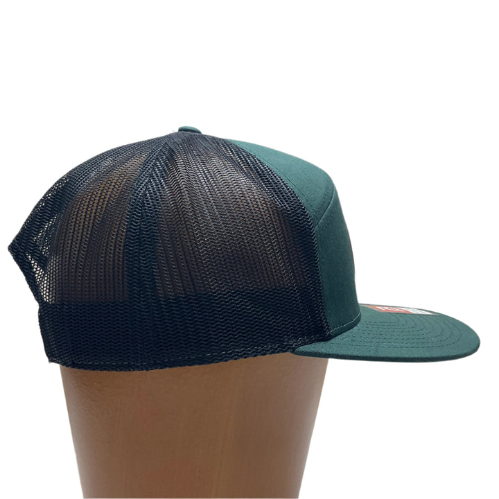 Side view of a Bigfoot Face Flexwood Patch Forest Green Flat Bill Trucker Hat, featuring a circular Flexwood patch etched with a Bigfoot face design and securely attached to a forest green and black Richardson flat bill trucker hat—handmade in Montana, USA by Last Best Supply Co.