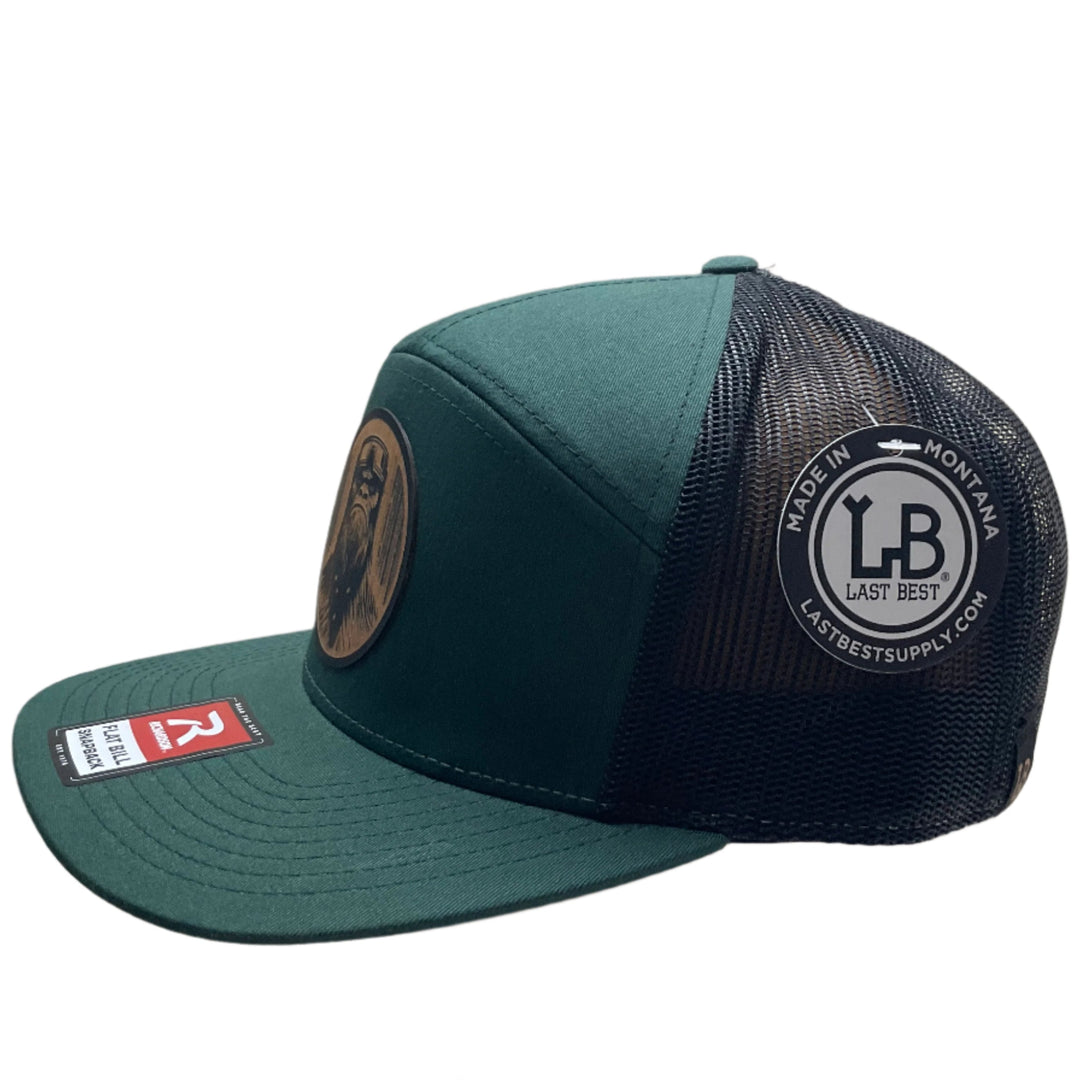 Side view of a Bigfoot Face Flexwood Patch Forest Green Flat Bill Trucker Hat, featuring a circular Flexwood patch etched with a Bigfoot face design and securely attached to a forest green and black Richardson flat bill trucker hat—handmade in Montana, USA by Last Best Supply Co.