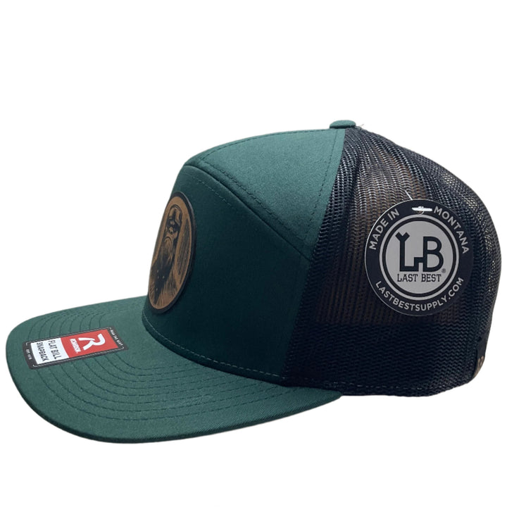 Side view of a Bigfoot Face Flexwood Patch Forest Green Flat Bill Trucker Hat, featuring a circular Flexwood patch etched with a Bigfoot face design and securely attached to a forest green and black Richardson flat bill trucker hat—handmade in Montana, USA by Last Best Supply Co.