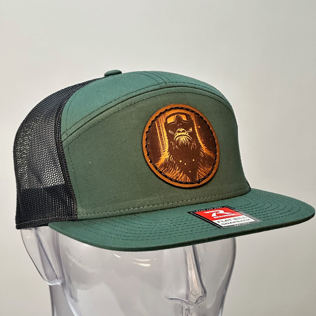 3/4 view of a Bigfoot Face Premium Leather Patch Forest Green Flat Bill Hat, featuring a circular hand-dyed leather patch etched with Bigfoot’s face, securely stitched with thick thread onto the structured front of a green and black Richardson flat bill trucker hat—handmade in Montana, USA by Last Best Supply Co.