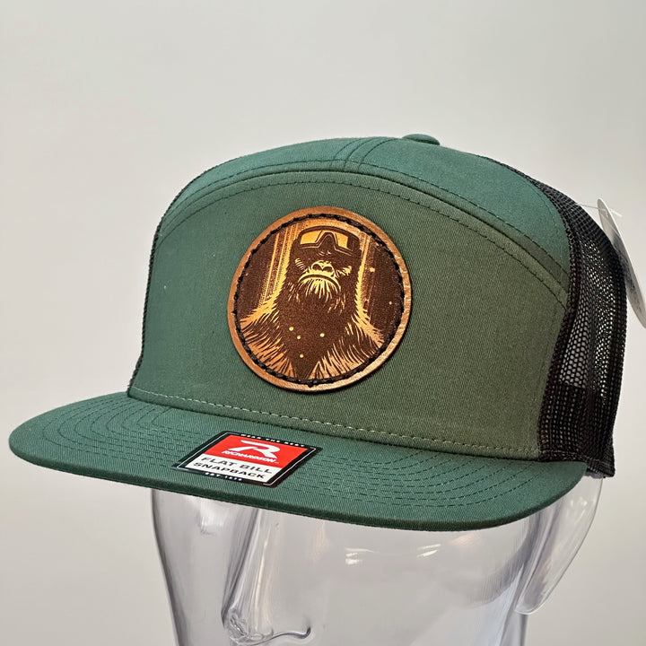 3/4 view of a Bigfoot Face Premium Leather Patch Forest Green Flat Bill Hat, featuring a circular hand-dyed leather patch etched with Bigfoot’s face, securely stitched with thick thread onto the structured front of a green and black Richardson flat bill trucker hat—handmade in Montana, USA by Last Best Supply Co.