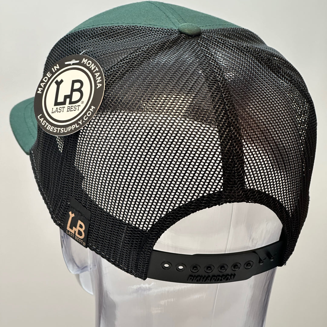 The back of a Bigfoot Face Premium Leather Patch Forest Green Flat Bill Hat, featuring a circular hand-dyed leather patch etched with Bigfoot’s face, securely stitched with thick thread onto the structured front of a green and black Richardson flat bill trucker hat—handmade in Montana, USA by Last Best Supply Co.