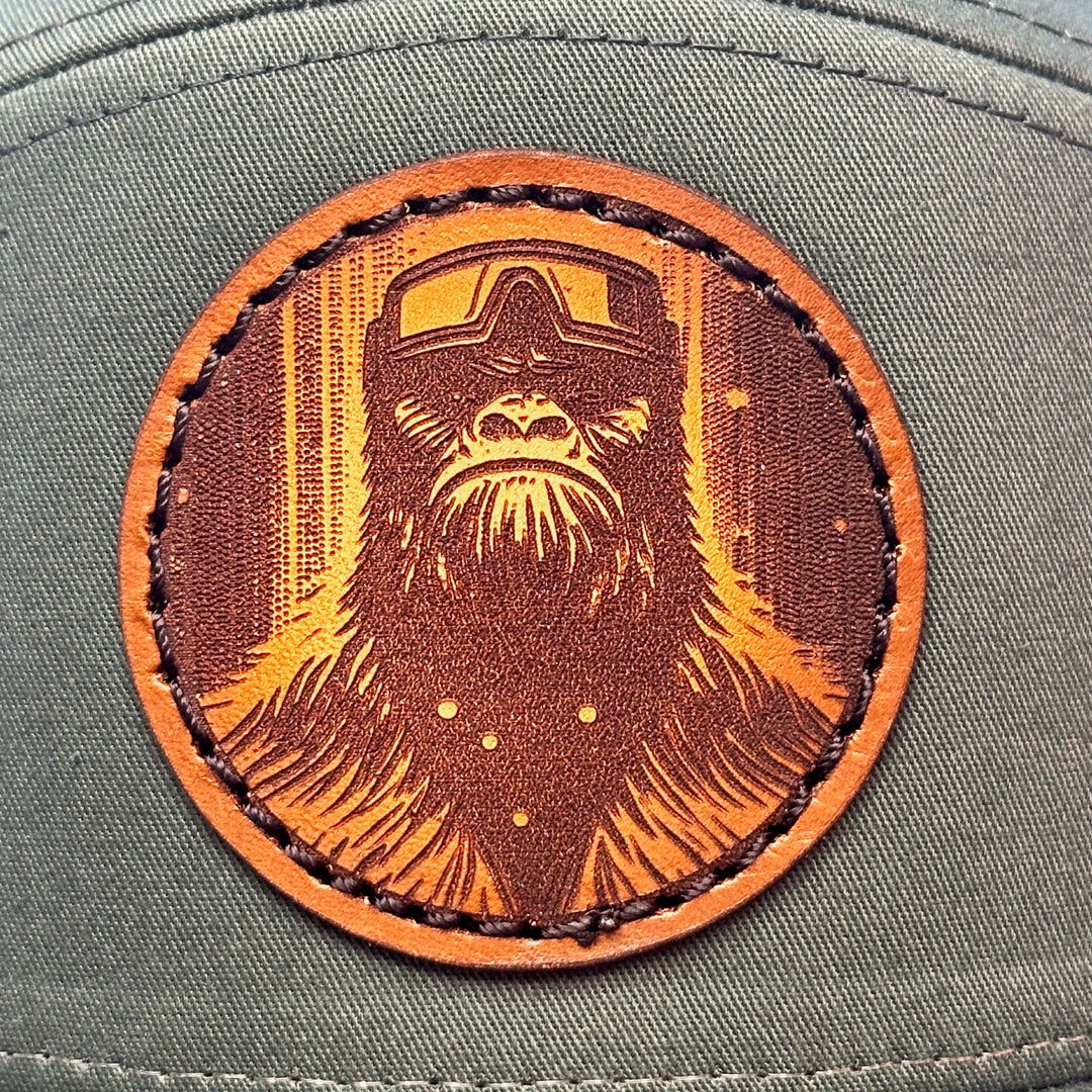 Patch detail of a Bigfoot Face Premium Leather Patch Forest Green Flat Bill Hat, featuring a circular hand-dyed leather patch etched with Bigfoot’s face, securely stitched with thick thread onto the structured front of a green and black Richardson flat bill trucker hat—handmade in Montana, USA by Last Best Supply Co.