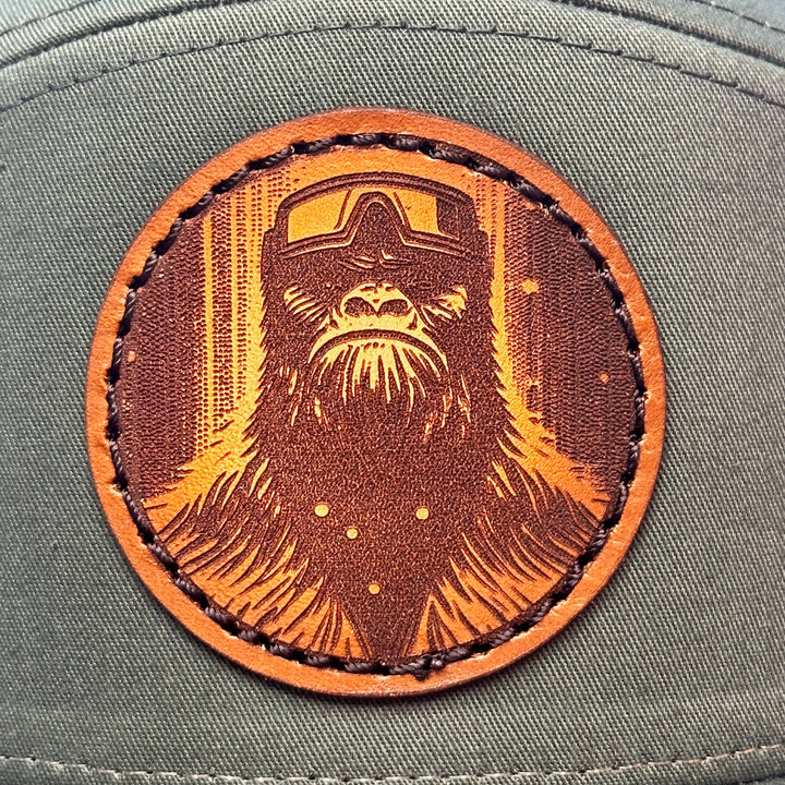 Patch detail of a Bigfoot Face Premium Leather Patch Forest Green Flat Bill Hat, featuring a circular hand-dyed leather patch etched with Bigfoot’s face, securely stitched with thick thread onto the structured front of a green and black Richardson flat bill trucker hat—handmade in Montana, USA by Last Best Supply Co.