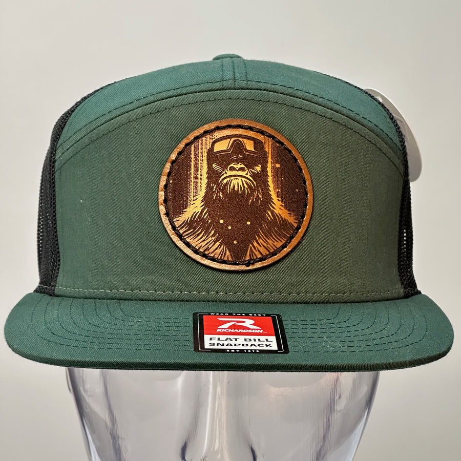 The front of a Bigfoot Face Premium Leather Patch Forest Green Flat Bill Hat, featuring a circular hand-dyed leather patch etched with Bigfoot’s face, securely stitched with thick thread onto the structured front of a green and black Richardson flat bill trucker hat—handmade in Montana, USA by Last Best Supply Co.