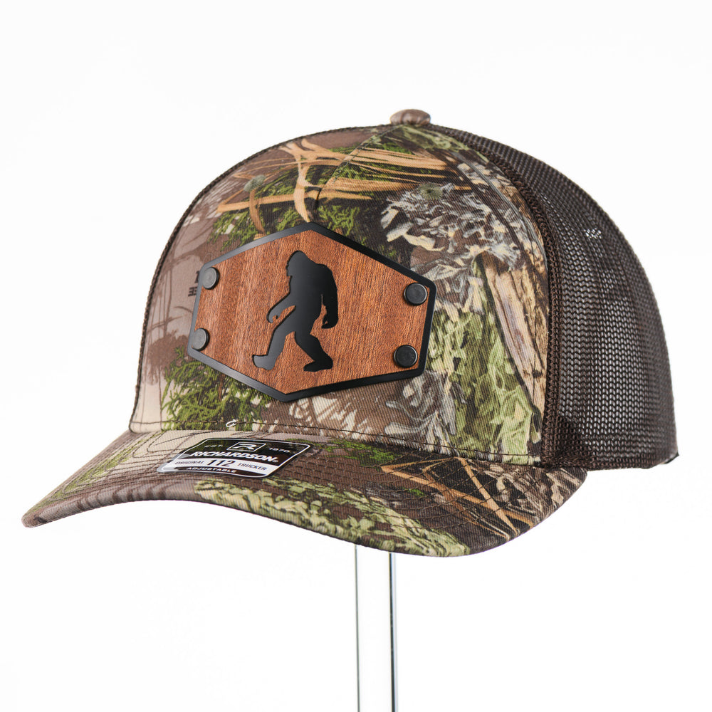3/4 view of a Bigfoot Mahogany Wood Hex Patch Camo Trucker Hat, featuring a hexagonal real mahogany wood veneer patch with a cutout silhouette of Bigfoot, laminated to a black anodized metal plate, and riveted to a camo and brown Richardson 112 trucker hat—handmade in Montana, USA by Last Best Supply Co.