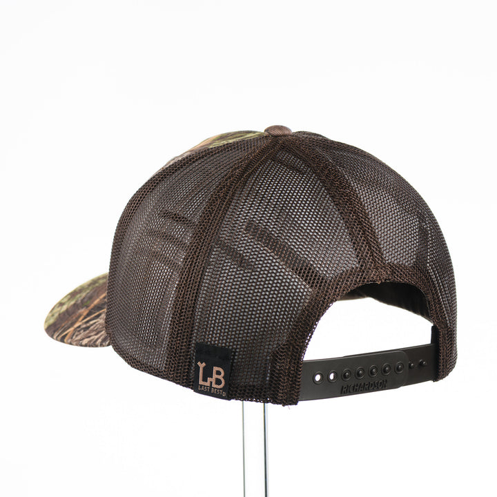 Back of a Bigfoot Mahogany Wood Hex Patch Camo Trucker Hat, featuring a hexagonal real mahogany wood veneer patch with a cutout silhouette of Bigfoot, laminated to a black anodized metal plate, and riveted to a camo and brown Richardson 112 trucker hat—handmade in Montana, USA by Last Best Supply Co.