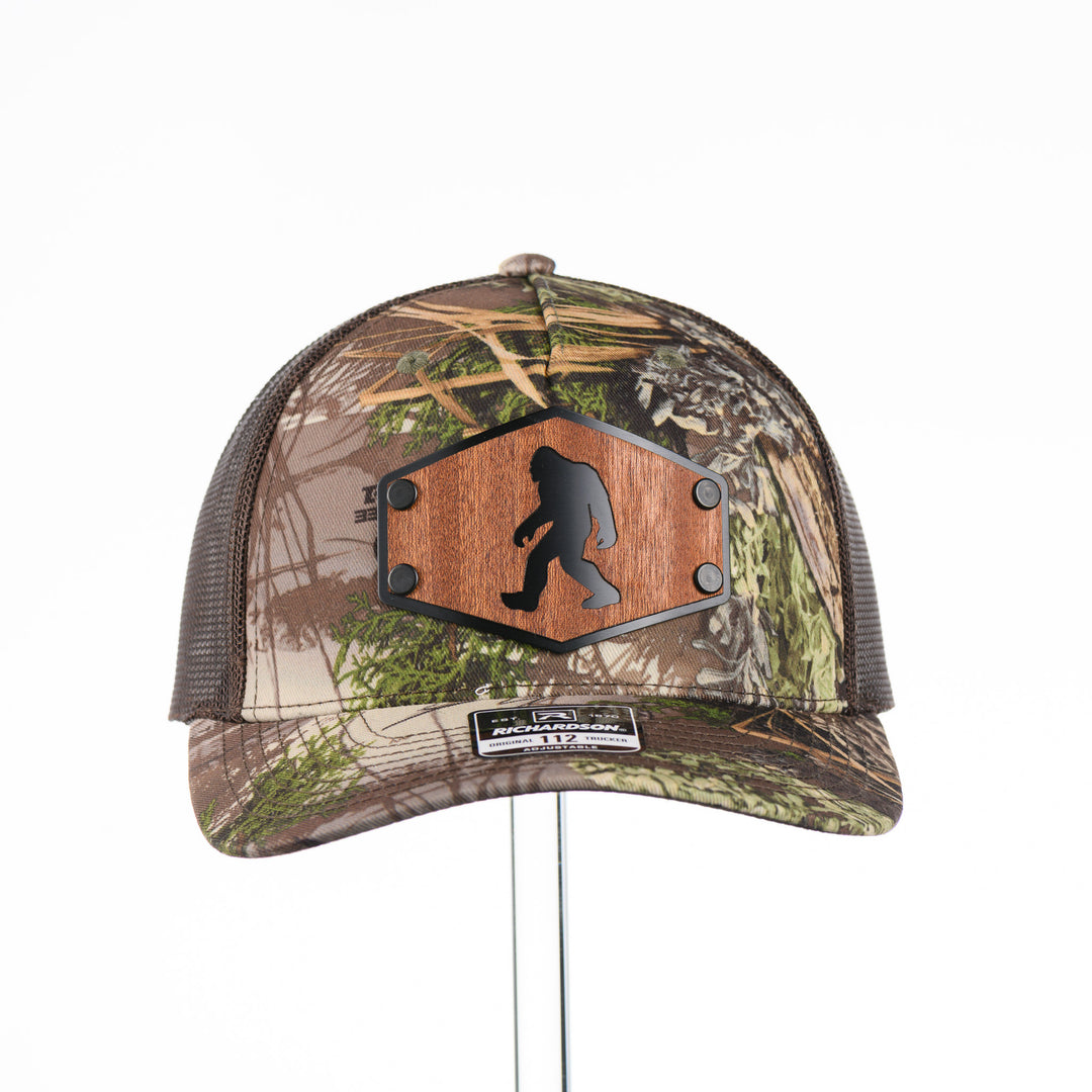 The front of a Bigfoot Mahogany Wood Hex Patch Camo Trucker Hat, featuring a hexagonal real mahogany wood veneer patch with a cutout silhouette of Bigfoot, laminated to a black anodized metal plate, and riveted to a camo and brown Richardson 112 trucker hat—handmade in Montana, USA by Last Best Supply Co.