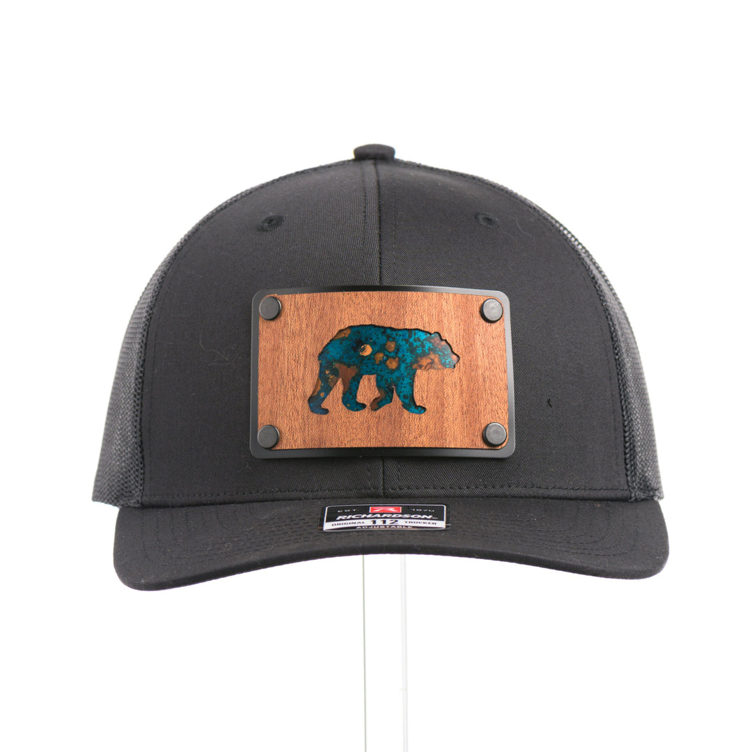 The front of a Black Grizzly Bear Teal Copper & Mahogany Wood Plate Patch Trucker Hat, featuring a rectangular real mahogany wood veneer patch with cut-out bear silhouette and teal copper inlay, laminated to a black anodized metal plate, and riveted to a black Richardson 112 trucker hat—handmade in Montana, USA by Last Best Supply Co.