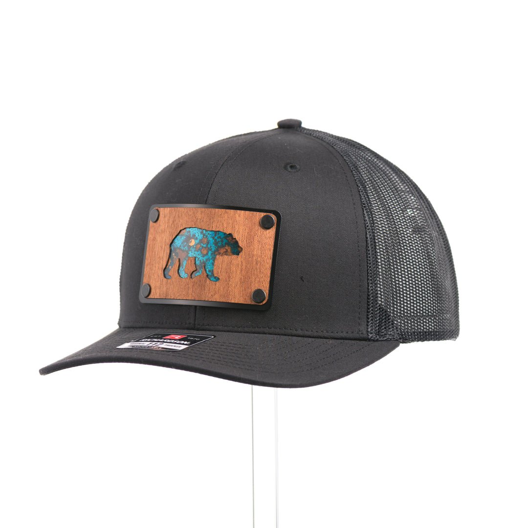 3/4 view of a Black Grizzly Bear Teal Copper & Mahogany Wood Plate Patch Trucker Hat, featuring a rectangular real mahogany wood veneer patch with cut-out bear silhouette and teal copper inlay, laminated to a black anodized metal plate, and riveted to a black Richardson 112 trucker hat—handmade in Montana, USA by Last Best Supply Co.