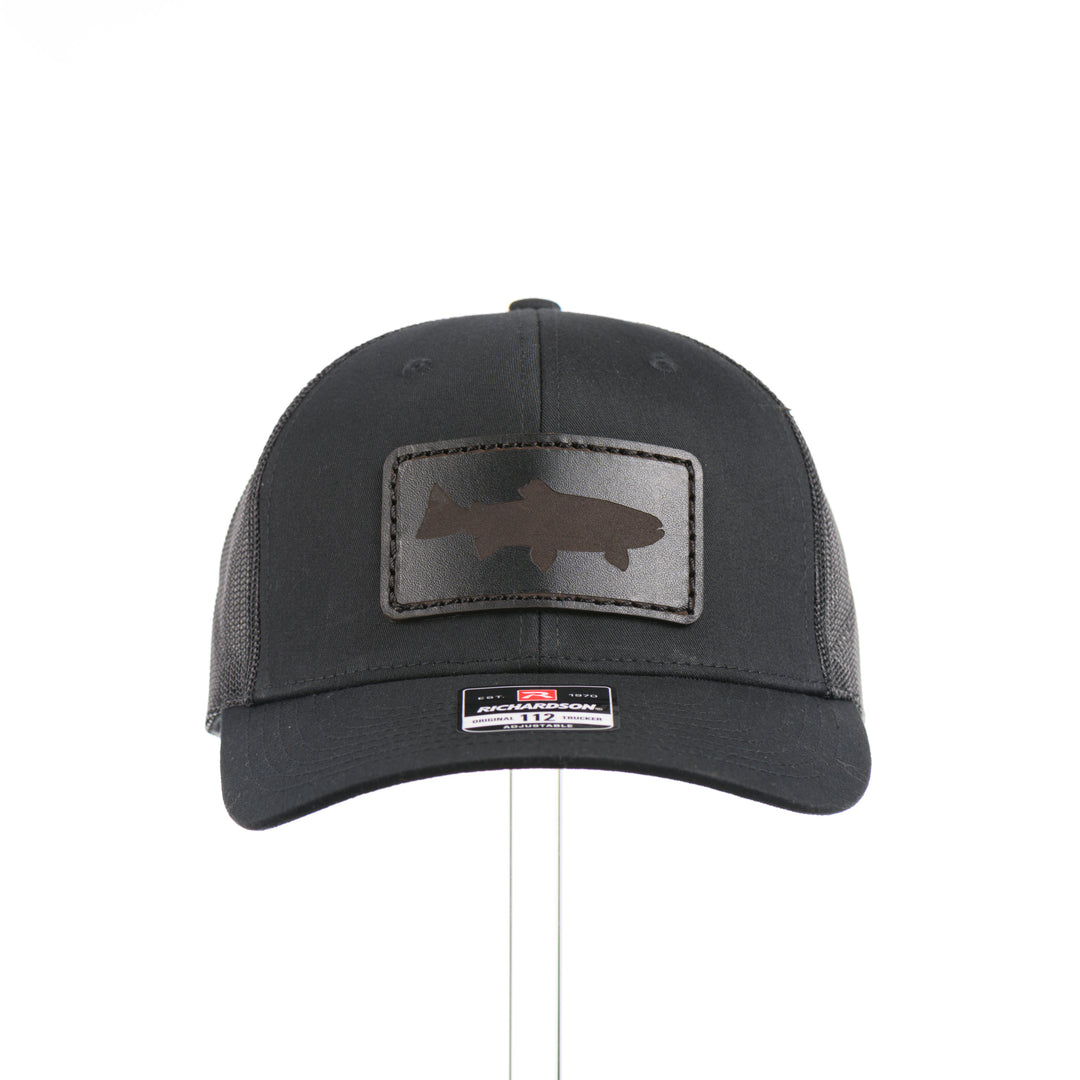 The front of an all black Richardson trucker hat with a black leather trout rectangle patch thick stitched on with black thread—handmade in Montana, USA by Last Best Supply Co.