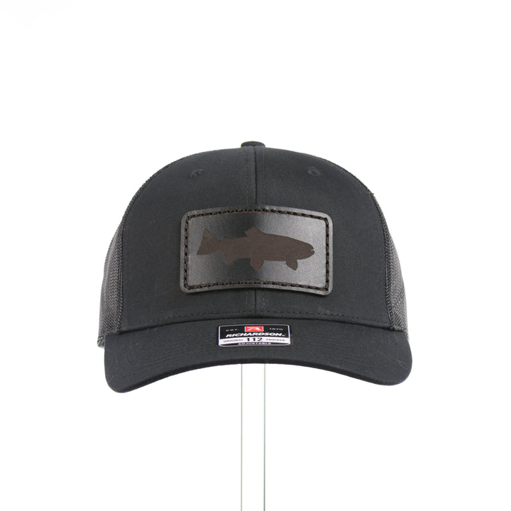 The front of an all black Richardson trucker hat with a black leather trout rectangle patch thick stitched on with black thread—handmade in Montana, USA by Last Best Supply Co.