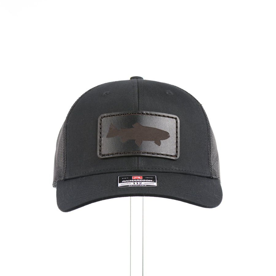 The front of an all black Richardson trucker hat with a black leather trout rectangle patch thick stitched on with black thread—handmade in Montana, USA by Last Best Supply Co.