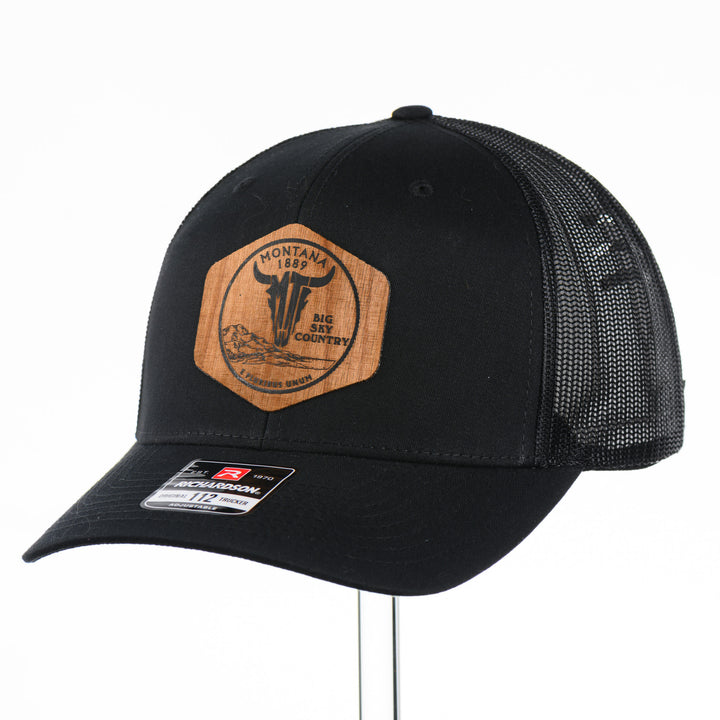 3/4 view of a Black Trucker Hat with Flexwood Patch Centennial Coin, featuring a hexagonal Flexwood veneer patch reverse etched with our iconic MT steer skull logo in a circle with the words "Montana 1889,” and "Big Sky Country," securely attached to a solid black trucker hat—handmade in Montana, USA by Last Best Supply Co.
