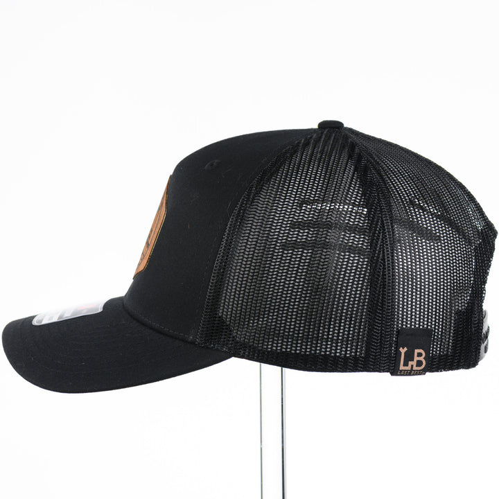 Side view of a Black Trucker Hat with Flexwood Patch Centennial Coin, featuring a hexagonal Flexwood veneer patch reverse etched with our iconic MT steer skull logo in a circle with the words "Montana 1889,” and "Big Sky Country," securely attached to a solid black trucker hat—handmade in Montana, USA by Last Best Supply Co.
