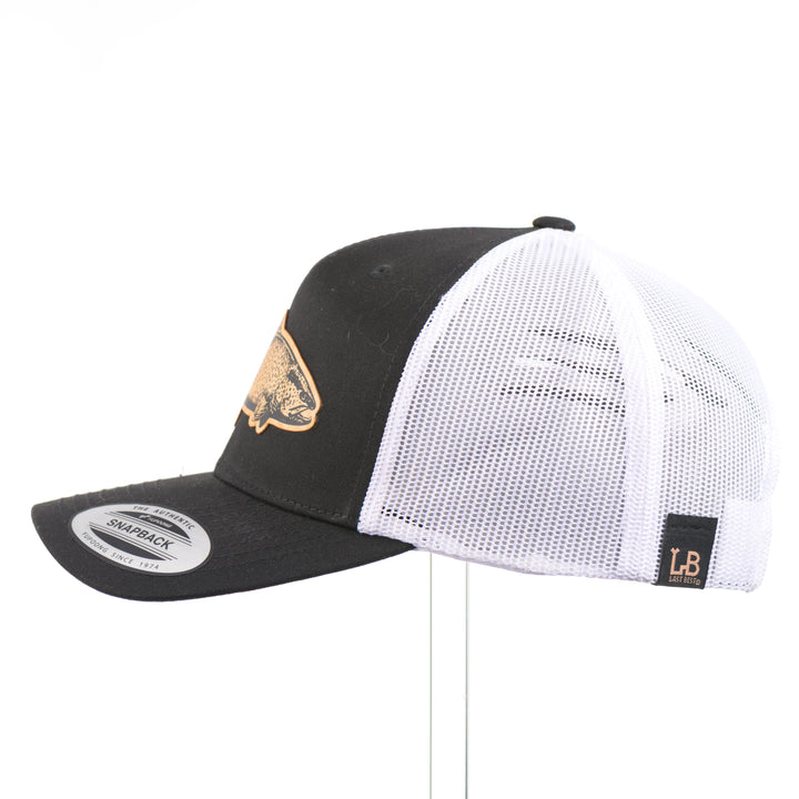 The side of a Sport-Tek Black & White Trucker Hat featuring a flexwood patch cut into the shape of a brown trout and etched with detail—handmade in Montana, USA by Last Best Supply Co.