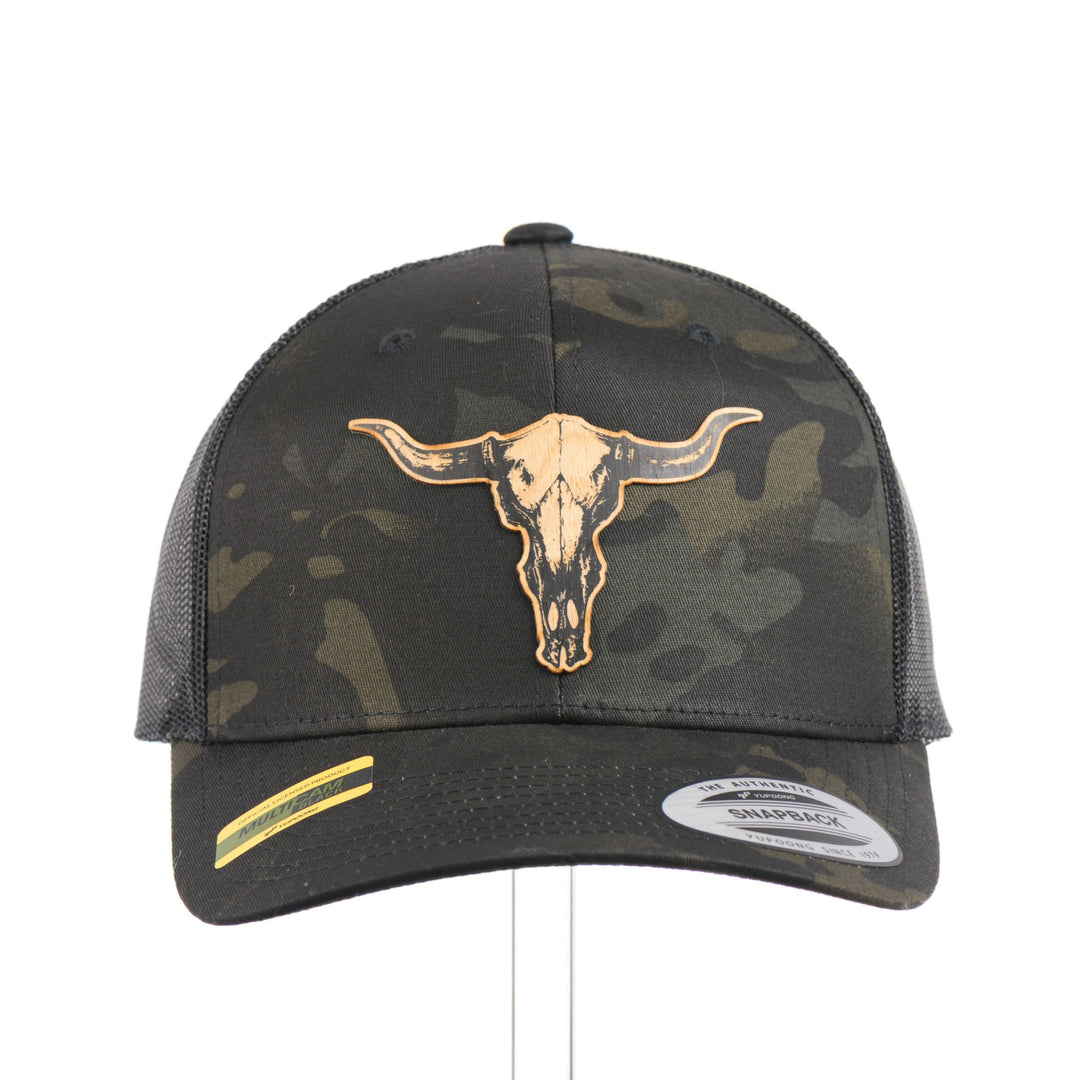 The front of a dark multicam trucker hat with a flexwood steer skull patch, handmade in Montana USA by Last Best Supply Co.