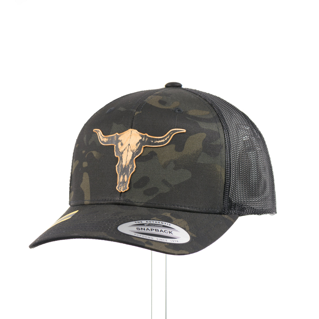 A dark multicam trucker hat with a flexwood steer skull patch at a quarter turn, handmade in Montana USA by Last Best Supply Co.